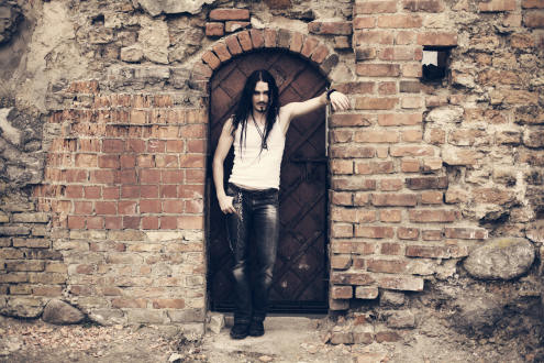 Kuvassa Nightwishin Tuomas Holopainen, mies jolla hihaton valkoinen paita yllään ja kuluneet farkut jalassa. Miehellä viikset ja leukaparta, pitkät mustat hiukset. Hän seisoo ränsistynyttä tiilimuuria vasten.