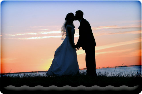Mies ja nainen suutelevat toisiaan meren rannalla kellertävässä auringonlaskussa. Naisella valkoinen häämekko ja miehellä musta puku. Maassa mustaa ruohikkoa.