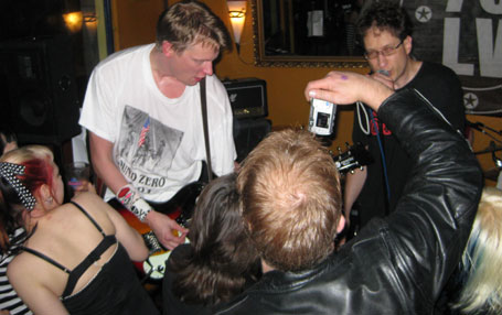 Valokuva Joey &amp; Tappajatomaatit -bändin jäsenistä, jotka soittavat lähietäisyydellä yleisöstä, joka on pakkautunut muusikkojen eteen. Kuvassa etualalla lyhythiuksinen mies ottaa valokuvaa. Taustalla näkyy kaksi muusikkoa soittamassa.