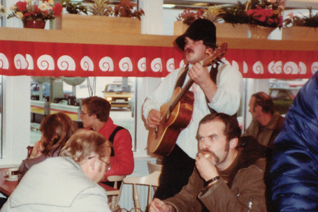 Vanha valokuva huopalakkipäisestä miehestä, joka soittaa akustista kitaraa baarissa tai kuppilassa. Miehellä viikset. Hänen ympärillään ruokailevia ihmisiä.
